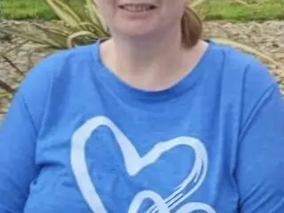 celticgoddessdom wears a blue t-shirt and smiles while sitting outdoors in a grassy area.