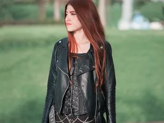 cherrryross wears a black leather jacket and corset top while standing outdoors in front of a green grass background.