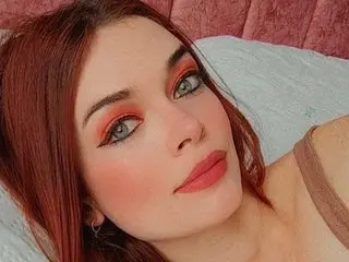 Young woman with red hair and warm sunset makeup relaxing on a white pillow in a cozy bedroom setting.