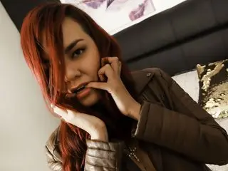 clairewitch poses with her hands near her face, showing her long red hair and wearing a brown jacket.