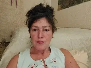 Middle-aged woman in a light blue floral sleeveless top relaxing on a white bed in a bright bedroom.
