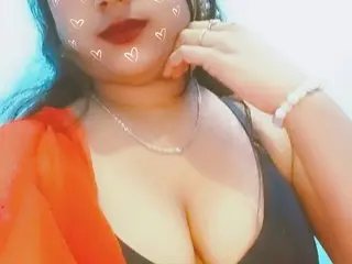 crazytara poses with her hands near her face, wearing a black tank top and showing her cleavage and long dark hair against a bright background.