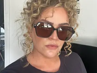 Blonde woman with curly hair wearing trendy dark sunglasses and a black t-shirt in a bright indoor setting.