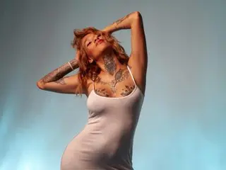 dakotastream poses with her hands behind her head, wearing a white tank top that shows her chest, neck, and arm tattoos against a plain background.
