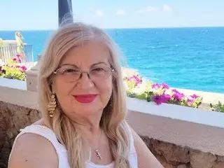 dariafloryss poses with long blonde hair and glasses, wearing a white tank top against a bright blue ocean background.