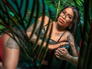 deborah_tatto is a tattooed woman wearing glasses and a black lace outfit, posing amidst green tropical leaves.