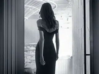 elena_wish stands with her back to the camera, showing her long dark hair and the curve of her back in a dark, form-fitting dress.
