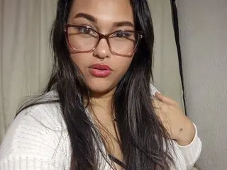 emma_miillerx poses close up wearing glasses and a white top, letting her long dark hair fall over her shoulders.