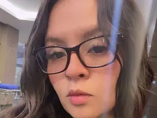 Close-up portrait of a young woman with long wavy hair wearing black-rimmed glasses in a soft indoor setting.
