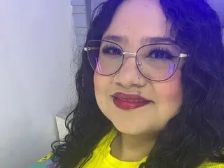 Smiling young woman with curly dark hair and glasses wearing a bright yellow shirt in a soft indoor setting.
