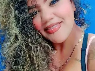 gisell_bigboobs smiles while posing with her long, curly blonde hair, showing her face and shoulders against a blue background.