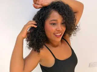 Smiling young woman with curly hair wearing a black tank top against a white wall.