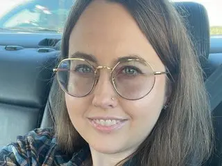 happylilcamgirl is a smiling woman with long brown hair and round glasses sitting in a car.