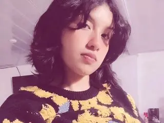 Young woman with dark wavy hair wearing a patterned yellow and black sweater in a soft indoor setting.