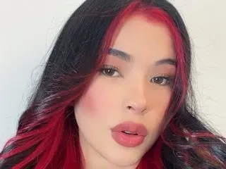 imnikaa poses close-up with long dark hair featuring bright red streaks, showing her face and shoulders against a plain white background.
