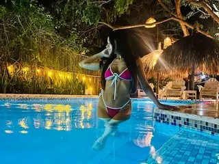 irinaalewis poses in a purple bikini while waist-deep in a swimming pool surrounded by tropical trees and dim lighting.