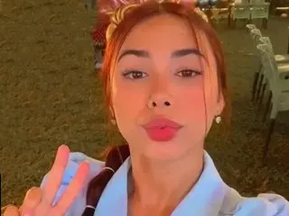 Young woman with reddish hair and light blue shirt making a peace sign in a warm outdoor setting.