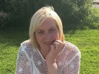jade50ff smiles while posing outdoors in a white lace top, showing her blonde hair and face against a green leafy background.