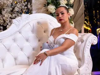 katiaexpensive poses on a white tufted sofa wearing a white off-the-shoulder dress with her hair pulled back.