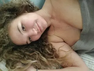 kayXXbaby smiles while lying down, showing her bare chest and long, curly brown hair.