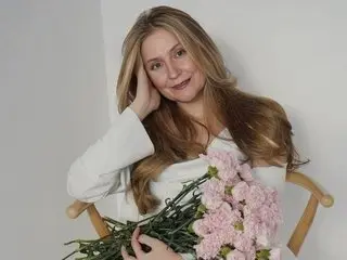 Smiling blonde woman in a white blouse holding pink flowers in a bright, minimalist indoor setting.