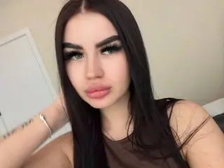 leonaמeilee has long dark hair and a soft expression, posing close-up in a brown top against a plain wall.