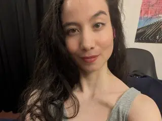 littleeveapples poses with long dark hair and a direct gaze, wearing a grey tank top against a dark background.