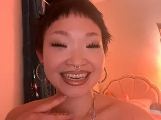 lolaspinner smiles, showing her teeth and a nose ring, with her short dark hair and silver earrings visible against a warm pink background.