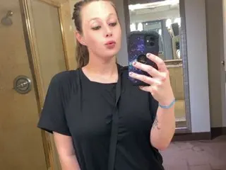 lovelyjane0699 poses for a mirror selfie wearing a black t-shirt, showing her face and hair pulled back in a bathroom setting.