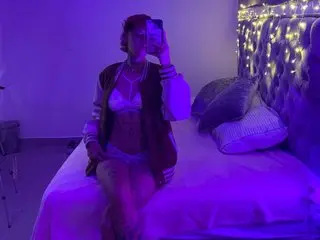 lucirosee poses on a bed wearing a white bra and dark bra, showing her cleavage and chest while looking at a phone in a purple-lit room.