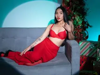 lucyliiuu poses on a grey sofa wearing a red bra and matching red skirt, showing off her tattoos and long dark hair against a blue background.