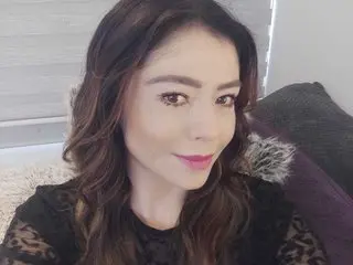 ludovicasstar poses with a soft smile, showing her long wavy brown hair and wearing a black lace top against a neutral background.