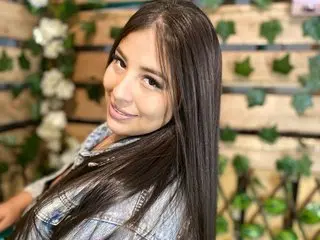 luisana_Milk has long, dark hair and a smile, posing in a denim shirt against a soft-focus background of green leaves and wooden slats.