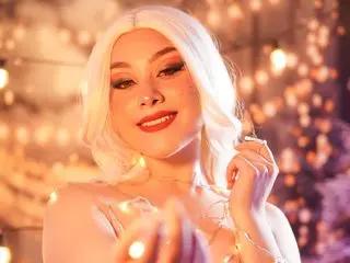 Glamorous woman with white hair and red lipstick posing amidst glowing fairy lights in a dreamy, warm setting.