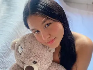 mia_celezte111 smiles while resting her chin near a soft teddy bear, showing her long dark hair and bare shoulders.