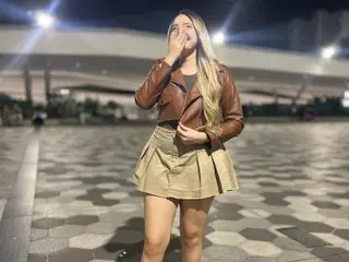 miarossexxXX wearing a brown leather jacket and tan skirt, posing outdoors with her hand near her mouth.