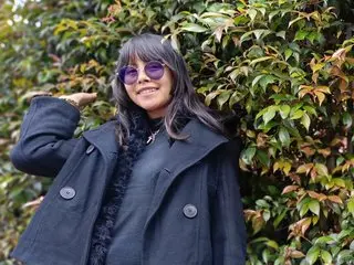 Smiling woman in black winter coat and purple sunglasses posing against lush green foliage.