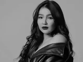 misssyoan poses with long dark hair wearing a black leather off-the-shoulder top against a plain grey background.