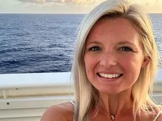 momixoxo smiles with her blonde hair down while posing outdoors near the blue ocean water.