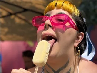 multiverseofmei wears pink sunglasses and licks a pale popsicle outdoors.