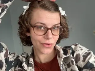 myprincessbliss has short wavy brown hair and wears glasses while posing in a patterned grey and white shirt.