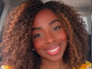 Smiling Black woman with voluminous curly brown hair and glowing makeup in a bright setting.
