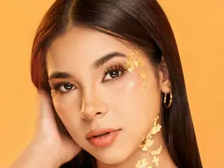 natalieheaven poses with long dark hair and gold leaf flakes applied to her face and neck against a solid orange background.