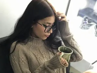 Thoughtful woman in a beige cable-knit sweater drinking tea in a bright, cozy indoor setting.