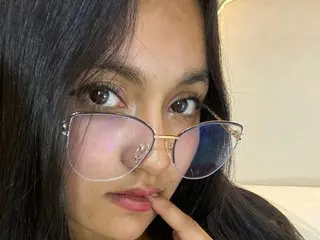 naughtyrose_vc — Young woman with long dark hair wearing oversized pink tinted glasses in a cozy indoor setting.