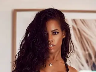 nicolleluv poses with long, wavy dark hair and a direct gaze, showing her bare shoulders and chest against a neutral background.