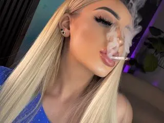 Glamorous blonde woman in a blue outfit exhaling smoke while smoking a cigarette in a moody indoor setting.