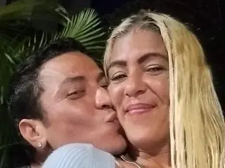 parrotclub69 with long blonde hair, smiling and kissing a man on the cheek in a dark setting.