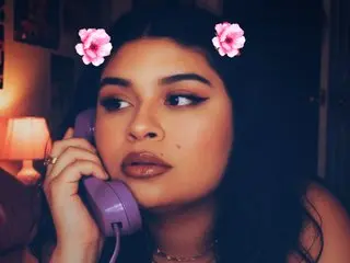 peacetorres18 poses with long dark hair, wearing a gold necklace and holding a purple telephone to her ear in a dimly lit room.