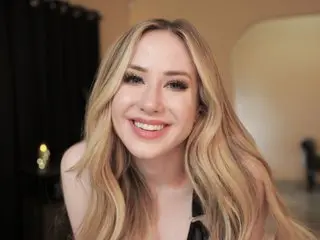 pintsizedblonde smiles while posing with her long blonde hair down, wearing a black top against a plain background.